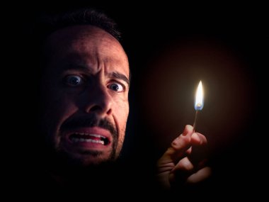 Karanlıkta elinde kibritle kameraya bakan endişeli bir adam. Karartma konsepti.