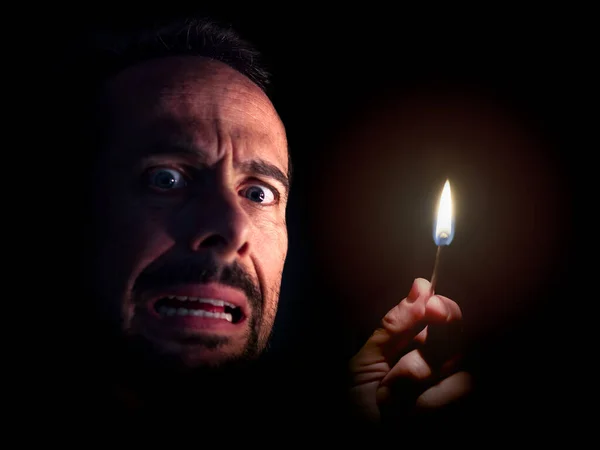 Worried man looking at the camera with a match in the dark. Blackout concept.