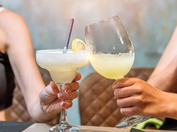 Two people holding cocktails, one margarita and one in a wine glass, garnished with lemon.