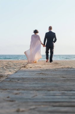 Resmi giyinmiş bir çift, arka planda deniz ve ufuk olan bir sahil kaldırımında el ele yürür..