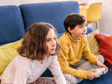 İki çocuk bir video oyununa dalmış, mavi bir kanepede oturuyor..