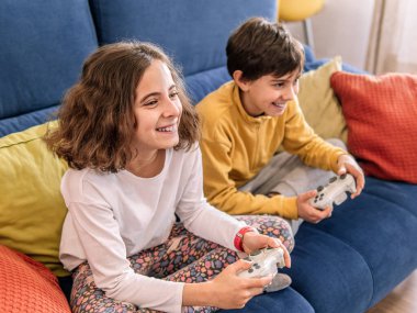 İki çocuk bir video oyununa dalmış, mavi bir kanepede oturuyor..