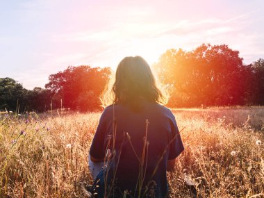 Bir çayırda oturan, gün batımına bakan, uzun otlar ve ağaçlarla çevrili, doğal huzurun tadını çıkaran bir birey..