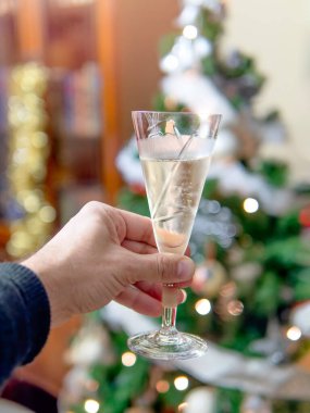 Arka planda Noel ağacı olan bir şampanya kadehini tutarken bir bayram kutlaması öneriliyor.