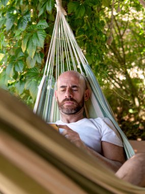 Kel adam hamakta eğleniyor. Etrafı yeşilliklerle çevrili. Açık havada akıllı telefon kullanırken gülümsüyor..