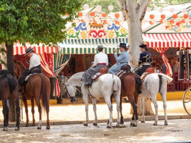 Seville, İspanya - 17 Nisan 2024: Arka planda şenlik dekorasyonları bulunan bir açık hava kültürel etkinliğinde geleneksel at sırtında İspanyol biniciler grubu.