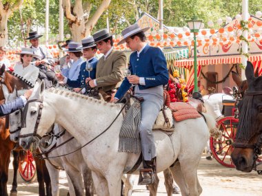 Seville, İspanya - 17 Nisan 2024: Arka planda şenlik dekorasyonları bulunan bir açık hava kültürel etkinliğinde geleneksel at sırtında İspanyol biniciler grubu.