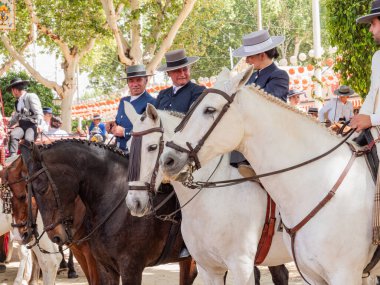 Seville, İspanya - 17 Nisan 2024: Arka planda şenlik dekorasyonları bulunan bir açık hava kültürel etkinliğinde geleneksel at sırtında İspanyol biniciler grubu.