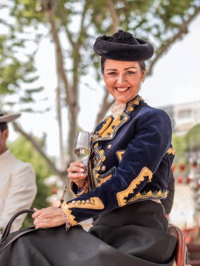 Seville, İspanya - 17 Nisan 2024: Geleneksel Endülüs kıyafetleri içinde zarif bir kadın, ata biniyor ve bir bardak şarapla Sevilla Fuarı 'nın tadını çıkarıyor..