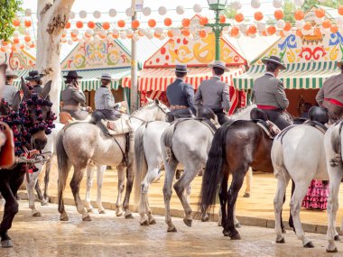 Seville, İspanya - 17 Nisan 2024: Arka planda şenlik dekorasyonları bulunan bir açık hava kültürel etkinliğinde geleneksel at sırtında İspanyol biniciler grubu.