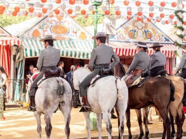 Seville, İspanya - 17 Nisan 2024: Arka planda şenlik dekorasyonları bulunan bir açık hava kültürel etkinliğinde geleneksel at sırtında İspanyol biniciler grubu.