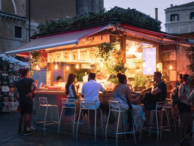 Bir Avrupa şehrindeki rahat, aydınlık bir sokak suyu barında sosyalleşen insanların akşam sahnesi, canlı bir atmosfer yaratıyor..