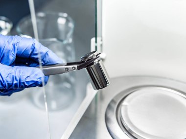 Bir operatörün eli analitik dengeyi sağlamak için çelik kalibrasyon ağırlığını tutuyor. Laboratuvarda kalite kontrolü kavramı.