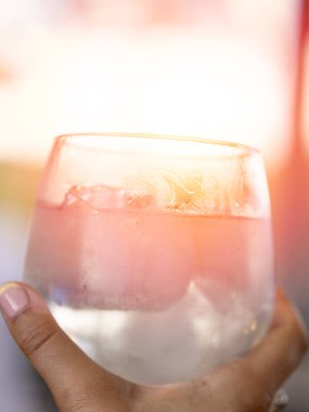 Bir el, buzlu kokteyl dolu buzlu bir bardağı tutar. Pembe bir eğilim ve yoğunlaşma içerir. Bulanık, güneşli bir arka plan üzerine kuruludur..