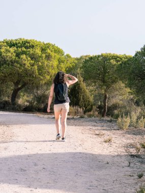 Sırt çantalı bir kadının arka görüntüsü, gözlerini koruyor ve güneşin aydınlattığı toprak yolda dikkatle bakıyor..