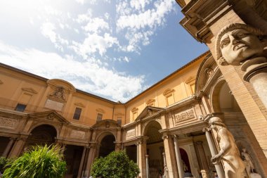 Vatikan 'daki Vatikan Müzeleri avlusunun güneşli manzarasında, parlak mavi gökyüzü altında Rönesans mimarisi ve klasik heykeller yer alıyor..