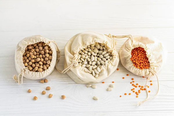 Open cotton bags with organic grains: lentils, chickpeas and beans on white wooden kitchen table. Top view