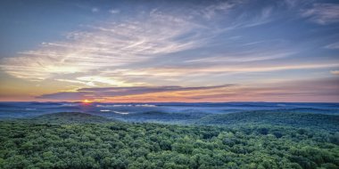 Hava manzarası, yemyeşil bir ormanın üzerinde canlı bir günbatımını yakalar ve dingin bir manzara yaratır..