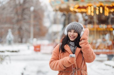 Pembe paltolu güzel, uzun saçlı kadın kış gününün tadını çıkar ve kar parkında kulaklıkla müzik dinle.