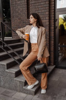 Korkulukların yanında poz veren, büyük ceketli, uzun boylu, şık bir kız ve şehrin arka planına basıyor.