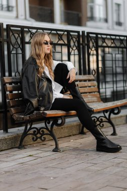 Şık, sarışın, siyah deri ceketli ve gözlüklü bir kadın şehirde bir bankta oturuyor. 
