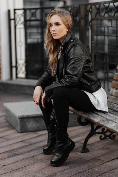 Fashionable  blonde woman model with  black leather jacket and style sunglasses sitting on a bench at the city