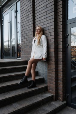 Sarışın, şık bir kadın, büyük boy beyaz ceketli ve elinde telefonla korkuluklara poz veriyor ve şehrin arka planında yürüyor.