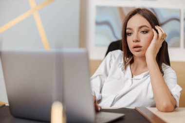 Business woman with long brunette hair looks down at her laptop screen at the office, hand on chin, seeming bored or uncertain
