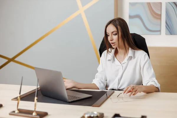 Smiling happy businesswoman sits at desk, looks at laptop screen, participating in self-improvement webinar, having fun on internet, communicating online by video call