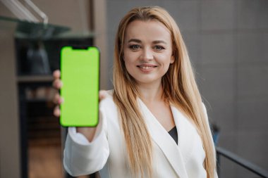 Büyük boy beyaz ceketli sarışın kadın şehir arka planında kromakey olan telefon ekranını gösteriyor.