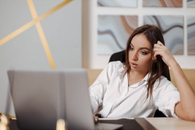 Business woman with long brunette hair looks down at her laptop screen at the office, hand on chin, seeming bored or uncertain