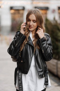 Beautiful smiling young blonde haired woman  in a black leather jacket using phone and making  call on a city street background