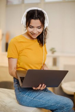 Close up portrait of indian or arabian woman inside home with headset for video call, woman smiling and looking at laptop, couch customer support tech helpline.