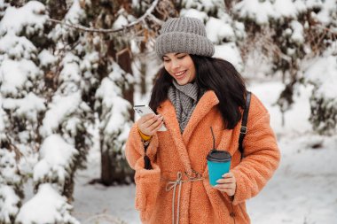 Pretty young long haired woman with amazing smile taking phone and a cup of coffee at a snowy winter park