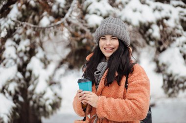 Güzel, uzun saçlı, harika gülüşlü bir kadın. Karlı bir kış parkında bir fincan kahve içiyor.