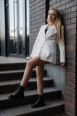 Blond stylish woman at oversized white jacket posing at the railing and steps on the city background