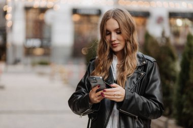 Güzel, gülümseyen, sarı saçlı, siyah deri ceketli bir kadın. Telefon kullanıyor ve şehrin arka planında video görüşmesi yapıyor.