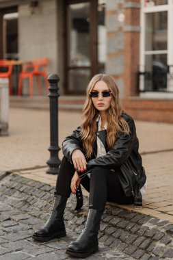 Fashionable  blonde woman model with  black leather jacket and style sunglasses sitting on a floor at the city 