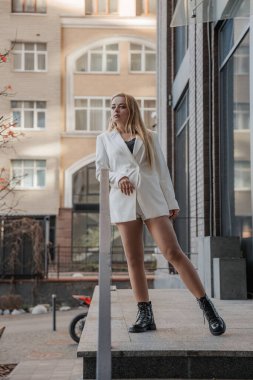 Blond stylish woman at oversized white jacket posing at the railing and steps on the city background