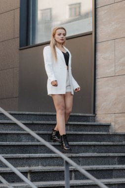 Blond stylish woman at oversized white jacket posing at the railing and steps on the city background