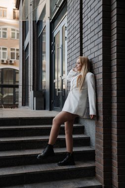 Blond stylish woman at oversized white jacket posing at the railing and steps on the city background