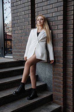 Blond stylish woman at oversized white jacket posing at the railing and steps on the city background