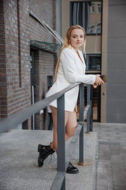 Blond stylish woman at oversized white jacket posing at the railing and steps on the city background