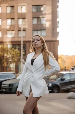 Blond stylish woman at oversized white jacket posing at the city background