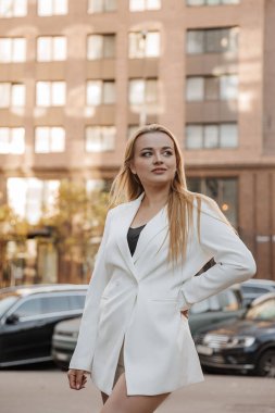 Blond stylish woman at oversized white jacket posing at the city background