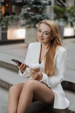 Büyük boy beyaz ceketli sarışın bir kadın telefon kullanıyor ve modern şehir arka planında aromalı kahve içiyor.