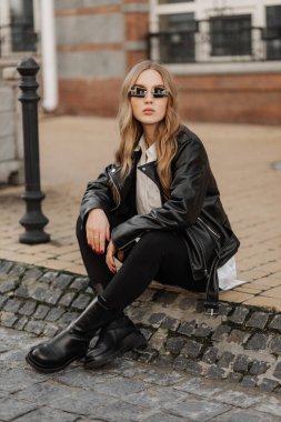 Fashionable  blonde woman model with  black leather jacket and style sunglasses sitting on a floor at the city 