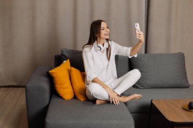 Kulaklıklı mutlu ve gülümseyen beyaz kadın cep telefonu kullanıyor. Evlerinde web kamerası dijital iletişimi var..