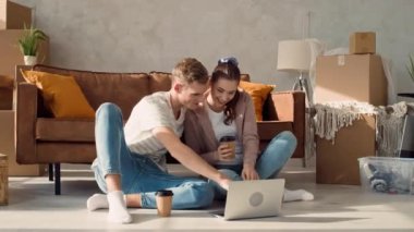 Couple in a New Home Using Laptop Drinking Coffee. Couple Using Laptop for Shopping, Booking Tickets, Discussing Interior. Girl Pointing in Screen.