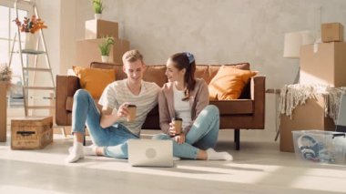Couple in a New Home Using Laptop Drinking Coffee. Couple Using Laptop for Shopping, Booking Tickets, Discussing Interior. Girl Pointing in Screen.